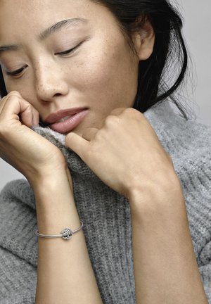 Bracciale d'argento con charm circolare esposto su una mano. Il modello indossa un maglione di lana grigio, evidenziando la texture della pelle e i toni naturali.