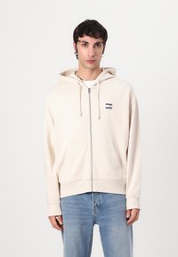 Beige lynlås hoodie med snore, frontlommer og et lille logo. Stoffet er blødt, med ribstrikkede manchetter og kant. Bæres over en hvid skjorte.