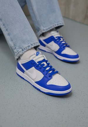 Białe i niebieskie buty sportowe Nike noszone z jasnoniebieskimi dżinsami i białymi skarpetkami na szarej plamistej podłodze.