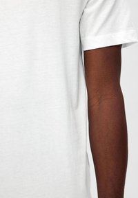 Camiseta de algodón blanca de mangas cortas, con un escote redondo y una textura suave, que cubre parcialmente un brazo de piel morena.