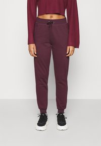 Pantalones de chándal color marrón con cintura elástica y cordón de ajuste, con piernas ajustadas y bolsillos laterales. Llevados con zapatillas negras.