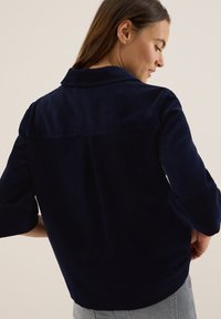 Vrouw met bruin haar in een marineblauwe corduroy jas en lichtgrijze broek, iets gedraaid om de details van de achterkant van de jas te tonen.