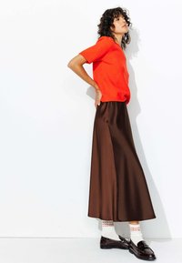 Un pull orange à manches courtes associé à une jupe maxi en satin marron. Agrémenté de chaussettes claires à motifs et de mocassins foncés.