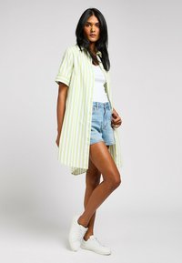 Maglietta a maniche corte verde e bianca a righe, tessuto leggero, orlo posteriore leggermente più lungo, abbinata a pantaloni corti di jeans e sneakers bianche.
