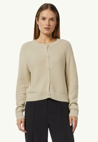 Beige knitted cardigan with round neckline, button closure, and ribbed cuffs. Features a cropped design and textured fabric.