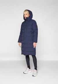 Jūras spēku garš pūkains mētelis ar horizontālu quilted dizainu, odere kapuci un priekšējo rāvējslēdzēju. Apvienots ar melniem legingiem un gaišiem sporta apaviem.