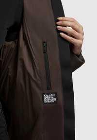 Brown jacket interior with black trim, featuring a zip pocket and a label that reads "Khujo™ original changing threads." Smooth texture.