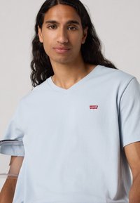 Lys blå V-hals t-skjorte laget av mykt stoff med korte ermer; har en rød Levi's-logo på venstre bryst. Jevn tekstur.