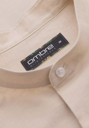Beige linnen shirtnek met een witte knoop en een label van stof met "ombre quintessence" in zwart, omrand met groene en bruine accenten.