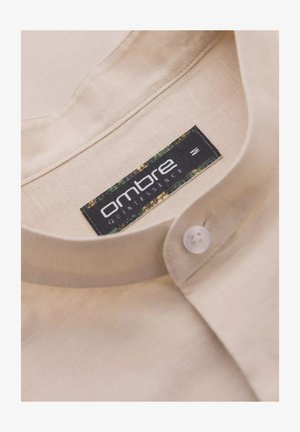 Beige linnen shirtnek met een witte knoop en een label van stof met "ombre quintessence" in zwart, omrand met groene en bruine accenten.