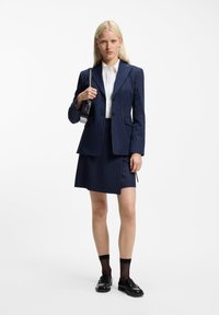 Marineblauw pinstripe pak met een op maat gemaakte blazer en rok, gecombineerd met een wit overhemd met knopen en zwarte loafers. Inclusief een kleine zwarte tas.