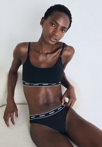 Woman with short hair wearing black Hugo branded bra and underwear, sitting on a textured beige chair against a plain white background.
