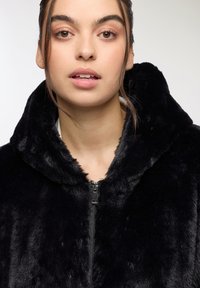 Giacca nera in pelliccia sintetica con cappuccio e chiusura a zip, caratterizzata da una texture morbida e un colletto spesso e accogliente. Design semplice senza dettagli hardware visibili.