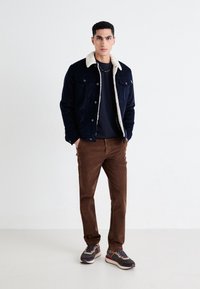 Chaqueta azul marino con un cuello de borrego color crema, camisa oscura debajo, pantalones de pana marrón y zapatillas deportivas multicolores.