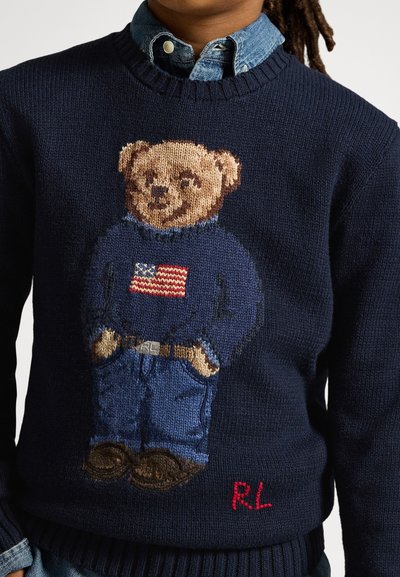 Marineblå genser med strikket bamse iført en genser med amerikansk flagg, jeans, brune sko og "RL"-initialer i rød søm.