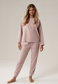 Kobieta stojąca boso, ubrana w jasnoróżową bluzkę z długim rękawem oraz spodnie z małymi dziurkami w całej powierzchni, na jednolitym tle.