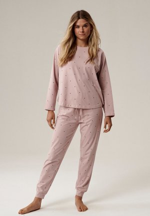 Kobieta stojąca boso, ubrana w jasnoróżową bluzkę z długim rękawem oraz spodnie z małymi dziurkami w całej powierzchni, na jednolitym tle.