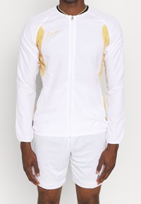 Athlet, der eine weiße, durchlöcherte Übergangsjacke mit gelben Seitenpaneelen und weiße Shorts mit schwarzem Logo am linken Bein trägt.