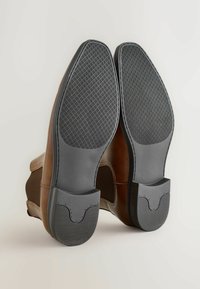 Zapatos de cuero marrón con suelas de goma negra que presentan un agarre texturizado y una entresuela suave. Punta redondeada y sutiles acentos de diseño en los laterales.
