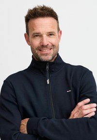 Chaqueta navy con cremallera y cuello alto, fabricada en tejido suave y con puños de canalé. Detalle de logo pequeño en el pecho en blanco y rojo.