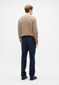 Beige, textúrájú pulóver, kerek nyakkivágással, sötétkék farmerrel párosítva, hátsó zsebekkel és barna bőrkiegészítővel a derékrészen. Fekete cipő.