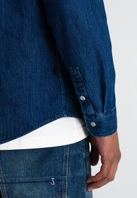 Denim overhemd in een donkerblauwe kleur, met knoopmanchetten en een gebogen zoom. GDragover een wit T-shirt, met een blauw textuurachtig materiaal op de achtergrond.