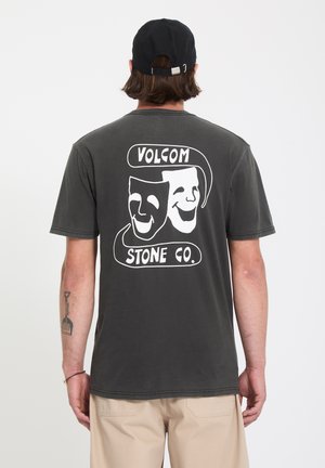 Personne portant un t-shirt Volcom Stone Co. gris foncé avec un motif de masques de comédie et de tragédie dans le dos, un pantalon beige et une casquette noire.