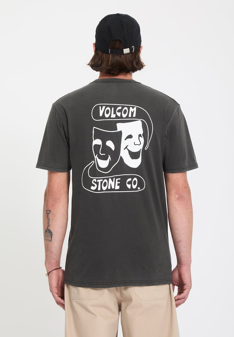 Άτομο που φοράει μπλούζα Volcom Stone Co. σε σκούρο γκρι με γραφικό από μάσκες κωμωδίας και τραγωδίας στην πλάτη, μπεζ παντελόνι και μαύρο καπέλο.
