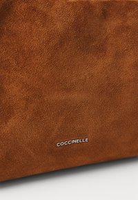 Brun ruskindshåndtaske med en blød tekstur, der har et sølvfarvet "Coccinelle" logo. Designet er enkelt og elegant, hvilket fremhæver sit håndværk.