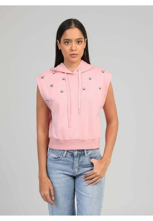 Femme portant un sweat à capuche sans manches rose avec des clous argentés, associée à un jean bleu clair, debout devant un fond gris uni.