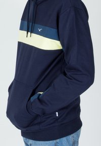 Persona che indossa un felpa blu navy con strisce orizzontali blu e gialle, mani nella tasca anteriore, abbinata a jeans blu.