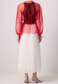 Rotes, durchsichtiges Oberteil mit langen Ärmeln, tailliert, kombiniert mit einem weißen, plissierten Rock. Das Outfit wird mit silbernen High Heels ergänzt.