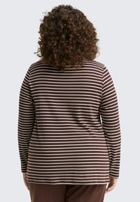 Person mit lockigem Haar, der ein langärmliges Oberteil in braun-weißen horizontalen Streifen trägt, von hinten vor einem schlichten Hintergrund gezeigt.