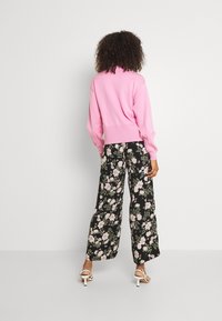 Pull rose tricoté avec poignets côtelés, associé à un pantalon large noir à motifs floraux. La tenue est complétée par des sandales blanches à talons.