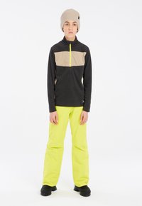Schwarzer Fleece-Pullover mit beigem Einsatz und gelbem Reißverschluss, kombiniert mit leuchtend gelben Hose. Das Outfit umfasst eine gestrickte beige Mütze und schwarze Stiefel.