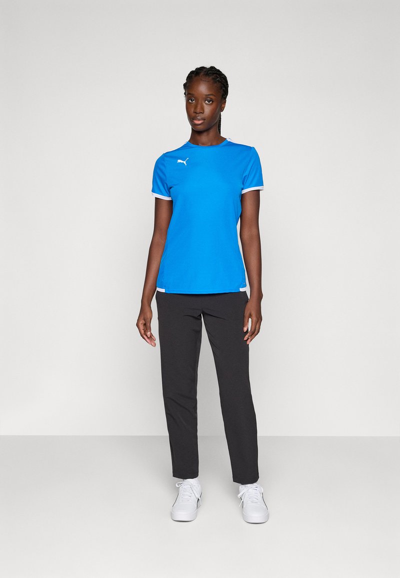 Blauw sportshirt met witte accenten en logo, gecombineerd met zwarte broek en witte sneakers. Model staat tegen een neutrale achtergrond.