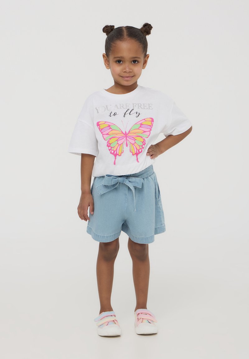 T-shirt bianco con una grafica di farfalla rosa e gialla, abbinato a pantaloni denim azzurri chiari con vita legata. Scarpe leggere con cinturini pastello.