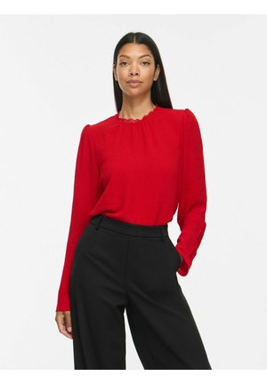 Femme aux longs cheveux foncés portant un chemisier rouge à manches longues et un pantalon noir taille haute, debout avec une main dans la poche sur fond blanc.