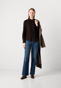 Woman wearing dark brown button-up shirt, blue wide-leg jeans, brown boots, holding brown coat over shoulder against plain background.