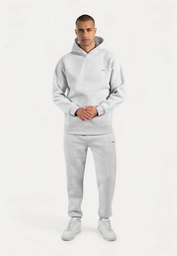 Grå hættetrøje med frontlomme og matchende joggingbukser. Fremstillet af blødt stof, har ribstrikkede manchetter og minimalistisk mærkning. Båret med hvide sneakers.