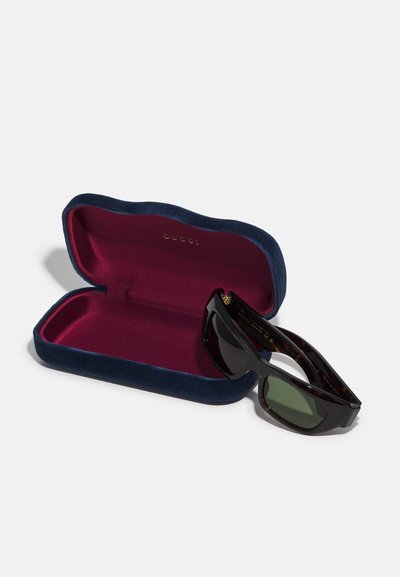 Occhiali da sole Gucci con montatura in tartaruga scura e lenti verde scuro, esposti all'interno di un astuccio in velluto blu navy con interno bordeaux.