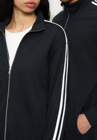 Δύο άτομα φορούν μαύρα μπουφάν με φερμουάρ και λευκές ρίγες στα μανίκια· το ένα έχει το χέρι στην τσέπη, μερική όψη των προσώπων και του κορμού.