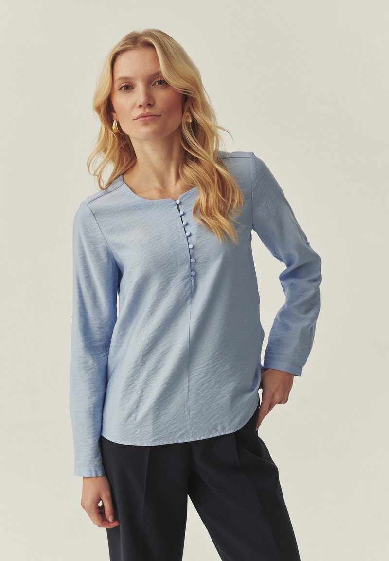 Blouse à manches longues bleu clair avec un col rond, un tissu texturé, et une rangée centrale de cinq boutons décoratifs. Portée avec un pantalon foncé.
