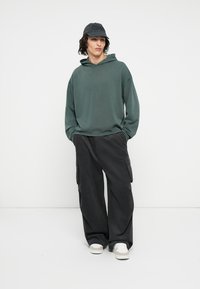 Mørkegrøn oversized hoodie med en frontlomme, parret med sorte cargo-bukser med sidelommer. Hvide sneakers fuldender outfittet.