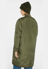 Grüne Nylon-Pufferjacke mit einem design, das bis zum Oberschenkel reicht, gerippten Bündchen und einer Reißverschlusstasche am linken Ärmel. Kombiniert mit einer gestrickten Mütze.