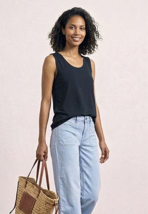 Kvinde med krøllet hår iført en sort ærmeløs top og lyseblå jeans, der holder en flettet stråtaske med brune læderstropper.