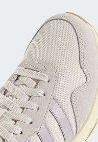 Detailní záběr špičky světle šedé tenisky se síťovinovým materiálem, semišovými prvky, odpovídajícími tkaničkami a světle levandulovými koženými pruhy na straně.