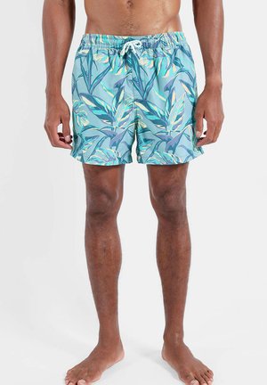 Homme portant un short de bain bleu clair avec un motif de feuilles tropicales vertes et violettes, debout pieds nus sur un fond uni.