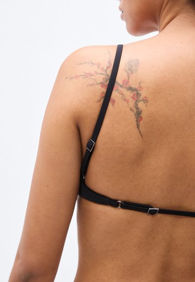 Μαύρο σουτιέν με λεπτές τιράντες και ασημένιο μεταλλικό ρυθμιστή, με τατουάζ λουλουδιού στον ώμο. Λείο υλικό και μινιμαλιστικός σχεδιασμός.