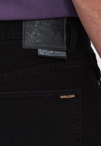 Jeans en denim noir avec une pièce en cuir texturé ornée d'un design en relief et du logo "VOLCOM" en doré sur la poche arrière, avec des accents cousus.